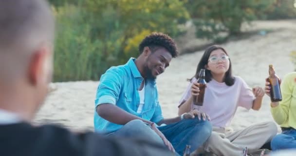 Un gruppo di amici è seduto sulla spiaggia vicino al lago, beve birra vicino al fuoco, canta canzoni, oscilla da un lato all'altro al ritmo della musica, persone di diverse nazionalità. — Video Stock