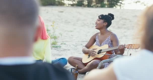 Une femme d'une beauté afro-américaine a les cheveux attachés dans un chignon joue de la guitare pour ses amis, les gens de différentes nationalités boivent de la bière au feu, chantent, dansent assis sur le sable au bord du lac — Video