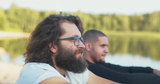 Zbliżenie na twarz dwóch mężczyzn siedzących nad jeziorem, słuchających muzyki przy ogniu, chłopiec z długimi włosami grzebie ją z powrotem, ma okulary, kołysze się do rytmu śpiewającego delikatnie — Wideo stockowe
