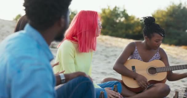 Skupina studentů tráví čas spolu sedí na pláži u jezera, zapalují táborák, pijí pivo ze skleněné láhve žena hraje na kytaru, zpívají, přátelská atmosféra — Stock video