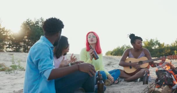 Un groupe d'étudiants passe du temps ensemble assis sur la plage près du lac, ils allument un feu de joie, boire de la bière de la bouteille en verre femme joue de la guitare, ils chantent, atmosphère amicale — Video