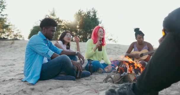 Un groupe d'étudiants passe du temps ensemble assis sur la plage près du lac, ils allument un feu de joie, boire de la bière de la bouteille en verre femme joue de la guitare, ils chantent, atmosphère amicale — Video