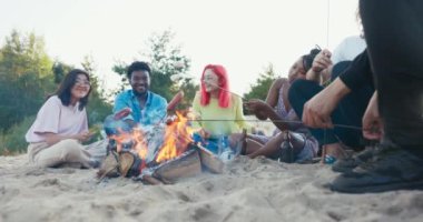 Farklı uluslardan arkadaşlar kumsalda ateşler içinde oturup bira içerek ateşte sosis kızartıyorlar lokum yiyip sohbet ediyorlar birlikte gülerek vakit geçirmeyi tartışıyorlar.