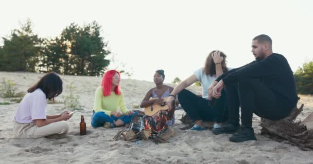 Przyjaciele siedzą przy ognisku na plaży, czekając, aż inni dołączą do nich pijąc piwo, śpiewając, bawiąc się. — Wideo stockowe
