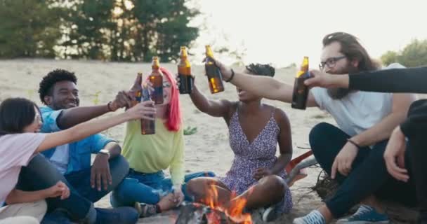Team von Freunden sitzt am See auf Sand, Strand die letzten Urlaubstage verbringen sie zusammen am Lagerfeuer, verschiedene Nationalitäten, trinken Bier in Flaschen zapfen sie jubelnd an — Stockvideo