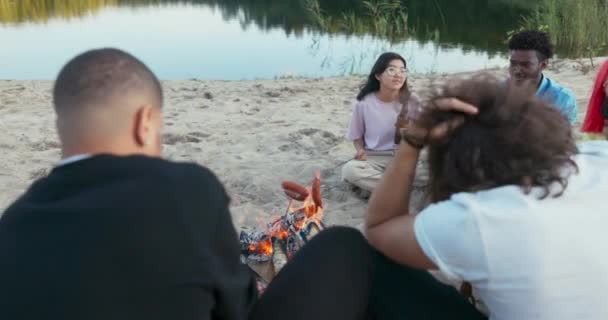 Piękna kobieta w okularach siedzi na piasku nad jeziorem, trzyma kiełbasę nad ogniskiem, smaży ją dokładnie po obu stronach, spędza czas z przyjaciółmi — Wideo stockowe