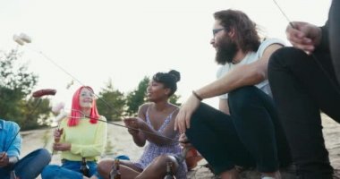 Bir grup iyi arkadaş dışarıda kamp ateşinin yanında, kumların üzerinde, ahşap kütüklerde, çubuklarla dolu sosisler ve marşmelovlar tutuyorlar, kızartıyorlar, mutlu, güler yüzlü, geveze insanlar.