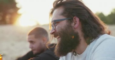 Profilde uzun saçlı ve kalın sakallı bir adamın yüzü görülüyor. Gözlüklü çocuk cam şişeden bira içiyor ve arka planda güneş ışınlarının altında arkadaşlarıyla konuşuyor.