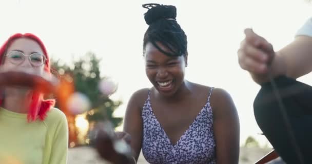 Smilende glad grinende kvinde med hendes hår bundet op i en bolle, sidder afrikansk-amerikansk kvinde på stranden med sine venner ved lejrbålet, steger pølser på pinde, fortæller vittigheder. – Stock-video