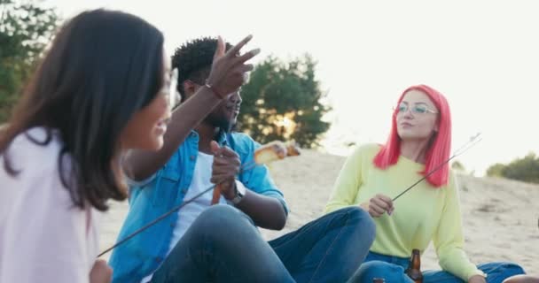 Přátelé sedí u letního táboráku ve večerních hodinách, slunce zapadá na pláži, odpočívají u zapáleného ohně, drží v rukou tyčinky s pečenými marshmallows a jí je žena olizuje špízu — Stock video