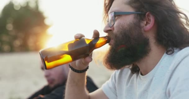 Mężczyzna z brodą i długimi włosami w okularach pije piwo ze szklanej butelki na tle zachodzącego słońca, spędzając czas z przyjaciółmi przy ognisku. — Wideo stockowe