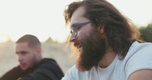 Das Gesicht eines Mannes mit langen Haaren und dickem Bart, der eine Brille trägt, ist im Profil zu sehen, Junge trinkt Bier aus Glasflasche und unterhält sich mit einem Freund, der draußen im Hintergrund der untergehenden Sonne sitzt — Stockvideo