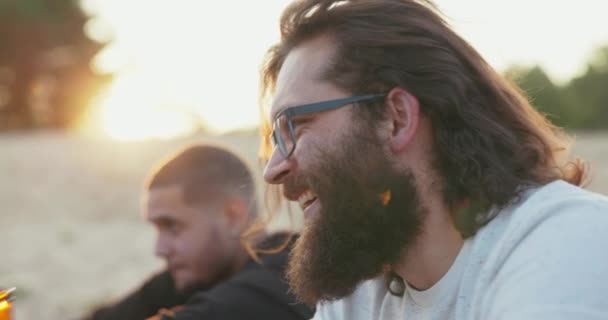 Das Gesicht eines Mannes mit langen Haaren und dickem Bart, der eine Brille trägt, ist im Profil zu sehen, Junge trinkt Bier aus Glasflasche und unterhält sich mit einem Freund, der draußen im Hintergrund der untergehenden Sonne sitzt — Stockvideo