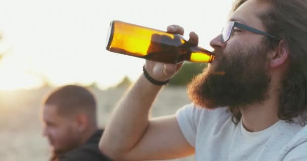 La cara de un hombre con el pelo largo y la barba gruesa con gafas es visible en el perfil chico está bebiendo cerveza de botella de vidrio y hablando con un amigo sentado fuera en los rayos de fondo de la puesta de sol resplandor — Vídeo de stock