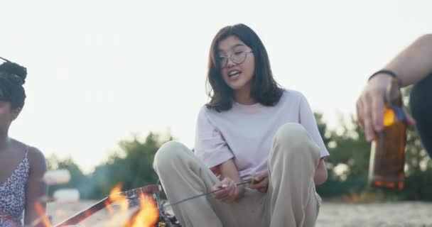 Uma mulher sorrindo modesta bonita com óculos A beleza asiática coreana está sentada perto do fogo ao lado de seus amigos, segurando nas mãos um pau no qual ela assa marshmallows. — Vídeo de Stock