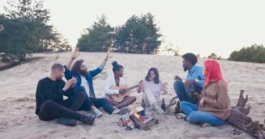 Arkadaşlar kumsalda birlikte eğleniyorlar kamp ateşinin yanında oturuyorlar, cam şişeler tutuyorlar bira içiyorlar el ele dans ediyorlar müzik eşliğinde şarkı söylüyorlar kadın gitar çalıyor