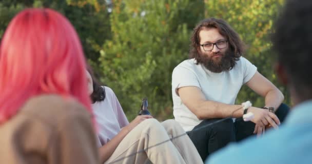 Mężczyzna z długimi włosami i brodą w okularach siedzi obok dziewczyny innej narodowości, pięknej kobiety azjatyckiej urody, rozmawia z przyjaciółmi, wymieniają opinie, piją razem piwo. — Wideo stockowe