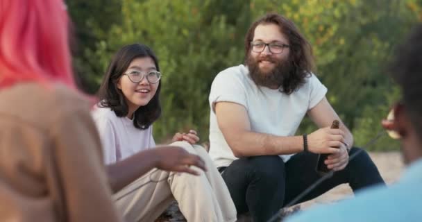 Egy férfi, hosszú hajjal és szakállal, szemüvegben ül egy másik nemzetiségű lány mellett, egy gyönyörű ázsiai szépséggel, beszélgetnek a barátokkal, véleménycserét folytatnak, együtt söröznek. — Stock videók