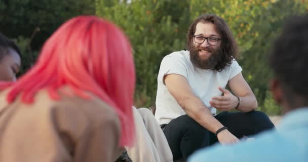 Ein Mann mit langen Haaren und Bart, der eine Brille trägt, sitzt neben einem Mädchen einer anderen Nationalität, einer schönen Frau asiatischer Schönheit, sie unterhalten sich mit Freunden, trinken zusammen Bier. — Stockvideo