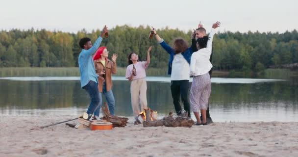 Studenci imprezują na plaży nad jeziorem, tańczą, śpiewają, bawią się przy zapalonym ognisku, trzymają butelki piwa, machają rękami w rytm muzyki — Wideo stockowe