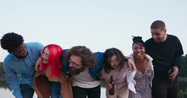 Een groep van beste vrienden van verschillende nationaliteiten omhelzen op een rij flessen in hun handen, zes glimlachende mensen leunen naar voren op hetzelfde moment kijken naar de camera, op de achtergrond een uitzicht op het meer — Stockvideo