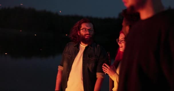 Gli amici si divertono di notte sul lago vicino al falò, le fiamme calde di legno li riscaldano, ballano, cantano, bevono birra, si divertono fino al mattino, festa degli studenti — Video Stock