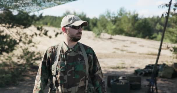 Soldato, militare, squadra di difesa territoriale uomo, vestito in abiti moro, occhiali neri si erge sullo sfondo della base, campo, incrocia le braccia sul petto, è fiducioso, coraggioso — Video Stock