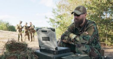 Askerler, ekipman silahlarıyla hazırlanmış, dışarıda bilgisayarın başında patronla görüntülü konuşma yapan kumandanı bekliyor.