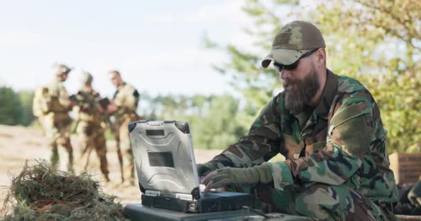 コンピュータに座って基地にデータを送信する司令官のために遠くに待っている機器や武器で準備され、服を着たフィールドでの行動に入るのを待っている兵士のグループ — ストック動画