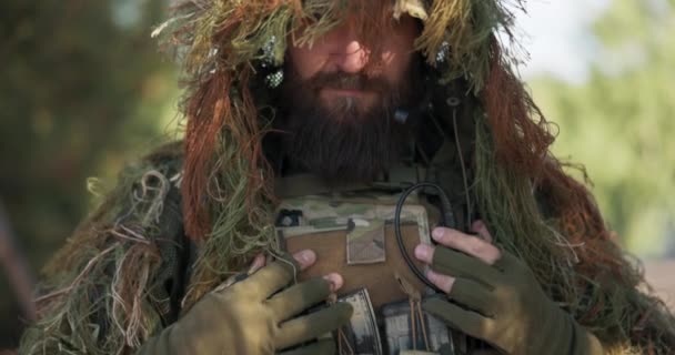 Poważny dzielny żołnierz ubrany w mundur wojskowy strzeże granic kraju, kryje się w lesie dzięki kamuflażowi na głowie trawy i mchu, na pasie piersiowym po akcesoria i sprzęt — Wideo stockowe