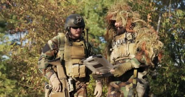 Dwóch żołnierzy przygotowanych do akcji, ubranych w mundur, kamuflaż omawiają strategię, topografię terenu patrząc na opancerzony laptop, planują trasę marszu wojsk przez las — Wideo stockowe