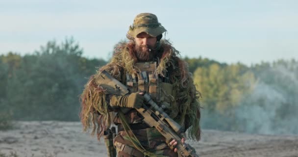 Portret van soldaat gekleed in camouflage uniform, helm en uitrusting gordel man staat in de aandacht met pistool in handen unholsters geweer, kijkt in de camera en heft wenkbrauwen als teken van uitdaging — Stockvideo