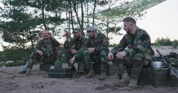 Katonák moro egyenruhában pihennek egy délutáni műszak után a bázison, kint ülnek a ládákon, tábortüzet készítenek, kolbászt pucolnak botokon, vacsorát készítenek. — Stock videók