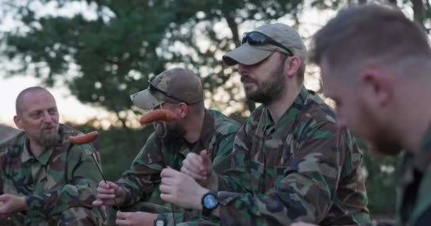 Военные в форме моро расслабляются после полуденной смены на базе в поле, сидят на ящиках снаружи и готовят костер, собирают колбасу на палочках, готовят обед, разговаривают, смеются — стоковое видео