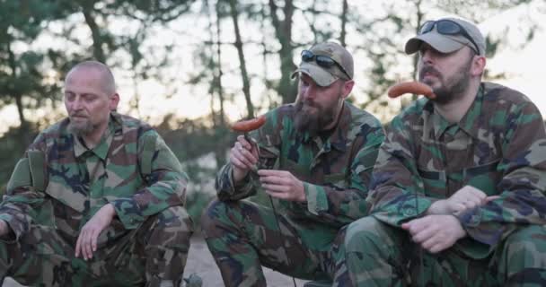 Military men in moro uniforms resting after an afternoon shift at base in field sitting on crates outside preparing campfire scooping sausage onto sticks guy with a beard cuts meat with a penknife — Stock Video