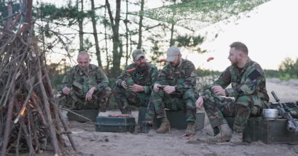 Katonák moro egyenruhában pihennek egy délutáni műszak után egy táborban, kint ülnek a ládákon és tábortüzet csinálnak, kolbászt lapátolnak botokra, alkoholt fogyasztanak. — Stock videók