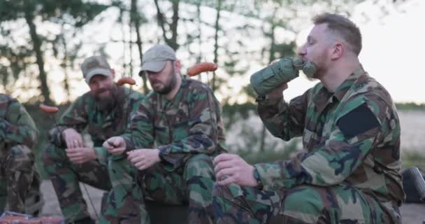 Vojenští muži v maskovacích uniformách odpočívají po odpolední směně na základně v poli, sedí venku na bednách a připravují táborák, dávají klobásy na tyčinky, pijí alkohol z láhve — Stock video