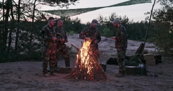 Τέσσερις άνδρες με στολές καμουφλάζ στέκονται γύρω από μια εστία με μια φωτιά μετά το ηλιοβασίλεμα, τηγανίζοντας λουκάνικα σε ξυλάκια, ένστολοι άντρες να ξεκουράζονται μετά το καθήκον, ασκήσεις πεδίου. — Αρχείο Βίντεο