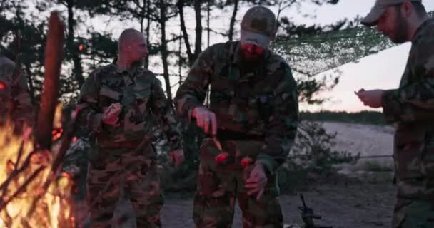 The soldiers are resting after their service in field they are warming themselves at hearth with bonfire, they are cutting sausages frying them on sticks, preparing supper, darkness falls after sunset — Stock Video