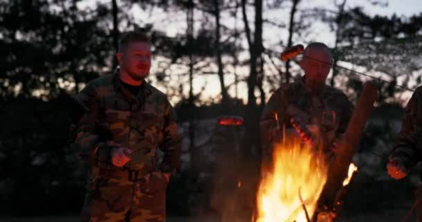 身着迷彩服的士兵们在谈话，他们在晚上讨论着用篝火煎香肠当晚餐，男人们在下班后休息、服侍、野外锻炼 — 图库视频影像