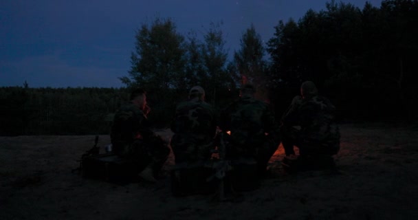 Un equipo de hombres en ropa de camuflaje está sentado en una colina en el campo por la noche, calentándose por una fogata encendida, freír salchichas para la cena, una excursión varonil, relajarse después del trabajo, servicio, caza — Vídeos de Stock