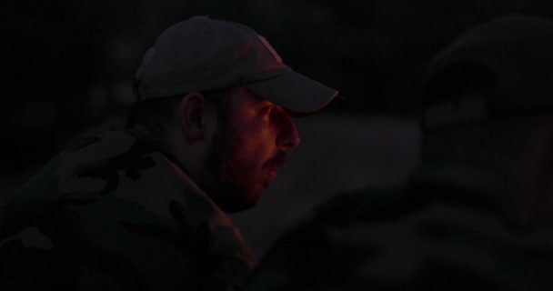 Gros plan d'un homme barbu visage en vêtements de camouflage et casquette la nuit éclairée par des flammes de feu de camp, il raconte une histoire effrayante à ses amis, camp de nuit, chasse — Video
