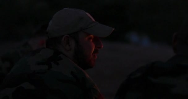 Close-up van een bebaarde man in camouflage kleren en pet 's nachts aangestoken door kampvuur vlammen, vertelt hij een eng verhaal aan zijn vrienden, nachtkamp, jagen — Stockvideo