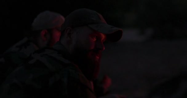 Detailní záběr obličeje vousatých mužů v maskovacích šatech a čepici v noci osvětlené ohněm, vypráví strašidelný příběh přátelům, svěřuje své problémy, noční tábor, lov — Stock video