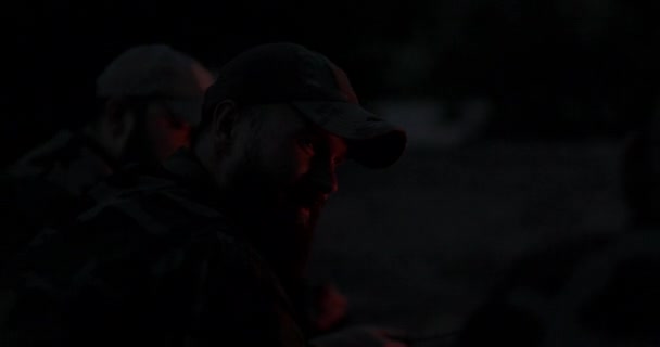 Detailní záběr na tvář muže s vousy v maskovacích šatech a čepici v noci osvětlené ohněm ohně, vypráví strašidelný příběh přátelům, vtipy, noční tábor, lov — Stock video