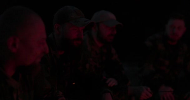 Un equipo de hombres en ropa de camuflaje está sentado en el campo por la noche, calentándose junto a una fogata encendida, freír salchichas para la cena, una excursión varonil, relajarse después del trabajo, servicio, caza — Vídeos de Stock