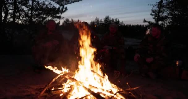 Uma equipe de homens em roupas de camuflagem está sentada em uma colina no campo à noite, aquecendo-se por uma fogueira acesa, fritando salsichas para o jantar, um passeio viril, relaxando depois do trabalho, serviço, caça — Vídeo de Stock