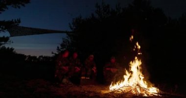 Büyük bir şenlik ateşi, arkasında askeri üniformalı adamlar gece şöminenin başında oturuyor, konuşuyor ve kendilerini ısıtıyorlar.