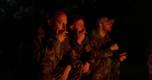Мужчины в камуфляжной одежде сидят у костра ночью, лицо освещено огнем, сидят на дежурстве, курят сигареты, разговаривают, рассказывают истории. — стоковое видео