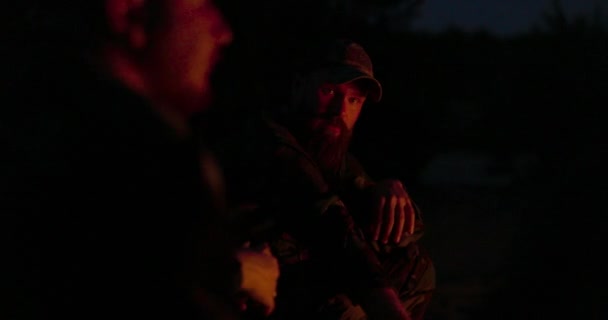 Mężczyźni ubrani w kamuflaż siedzą nocą przy ognisku, rozpaleni ogniem, siedząc na służbie, paląc papierosy, rozmawiając, opowiadając historie. — Wideo stockowe